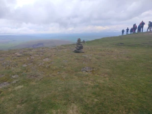Kopiec kamieni na szczycie wzgórza, a w tle widoczne sylwetki turystów. Ten surowy pejzaż skłania do zadumy nad siłą gór, które potrafią jednocześnie onieśmielać i zapraszać, przypominając nam o pielgrzymce w głąb samego siebie, jaką podejmujemy, wędrując pośród natury Pen-y-ghent, Anglia. Fot. Piotr Biegasiewicz
